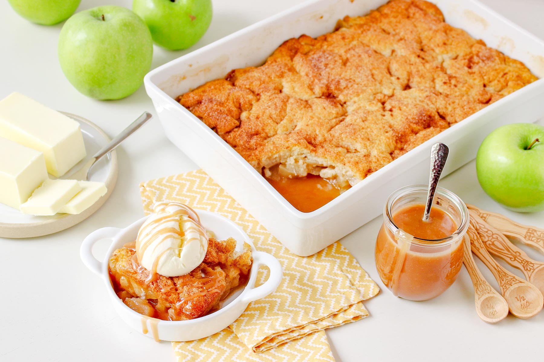 apple cobbler in large white baking dish with caramel sauce jar off to the side and green granny smith apples next to it. serving in small white bowl topping with ice cream and caramel sauce.