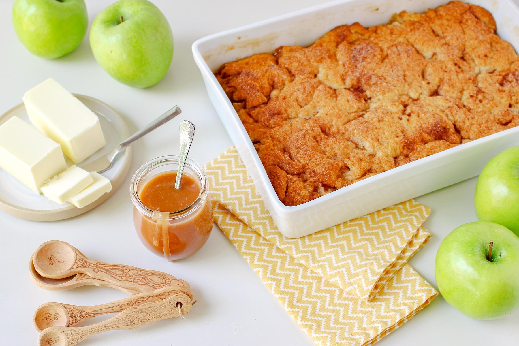 apple cobbler in large white baking dish with caramel sauce jar off to the side and green granny smith apples next to it.