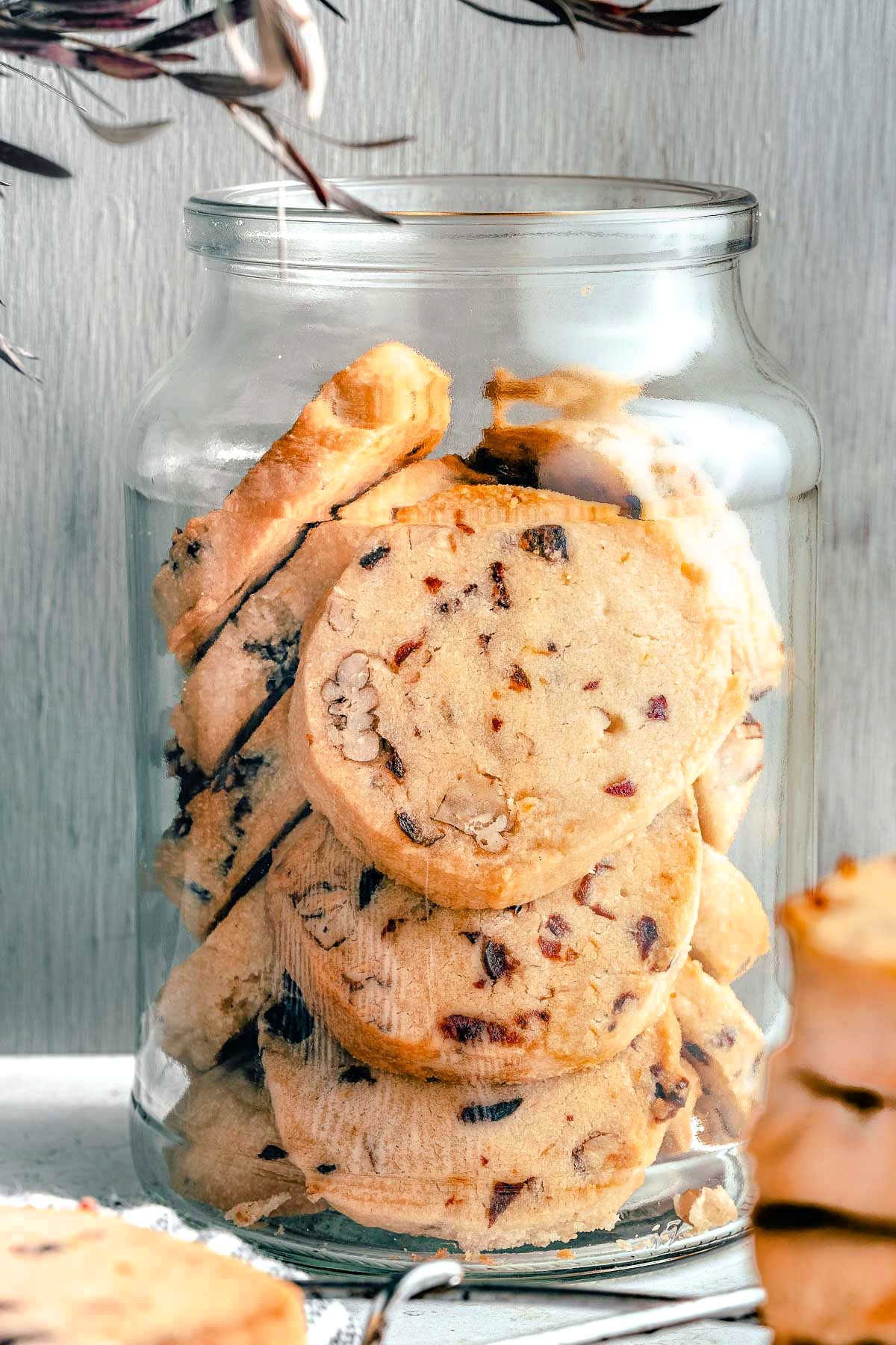 glass jar full of cranberry shortbread cookies. stack of cookies sitting in front of jar.
