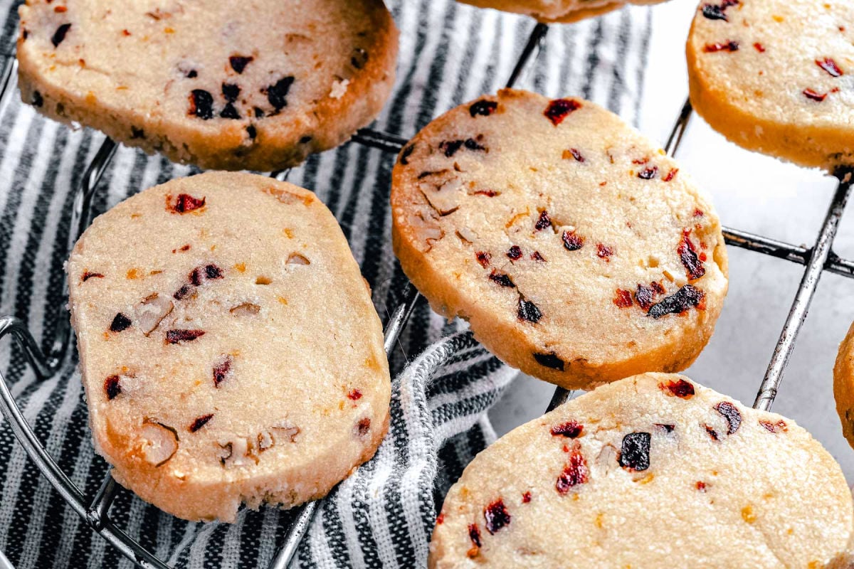 five shortbread cookies with cranberries and pecans on wire rack next to striped towel.