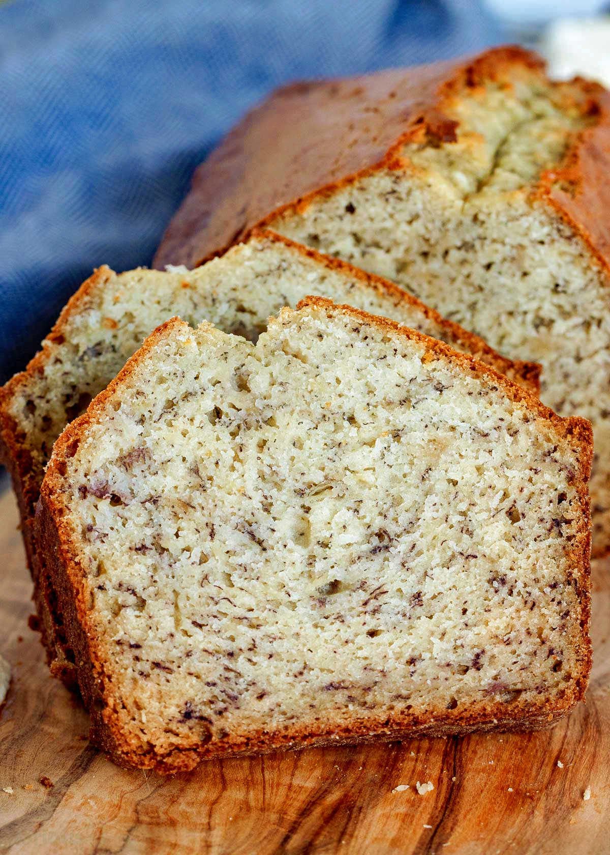 two slices of banana bread resting against the rest of the loaf in front of a blue napkin and sitting on a wood board.