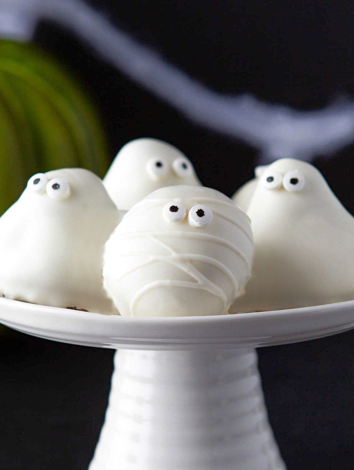 mummy and ghost oreo balls on white cake stand with green pumpkin in background.