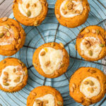 eight pumpkin muffins with cream cheese fillings are sitting on a wire rack on top of a light aqua colored napkin.