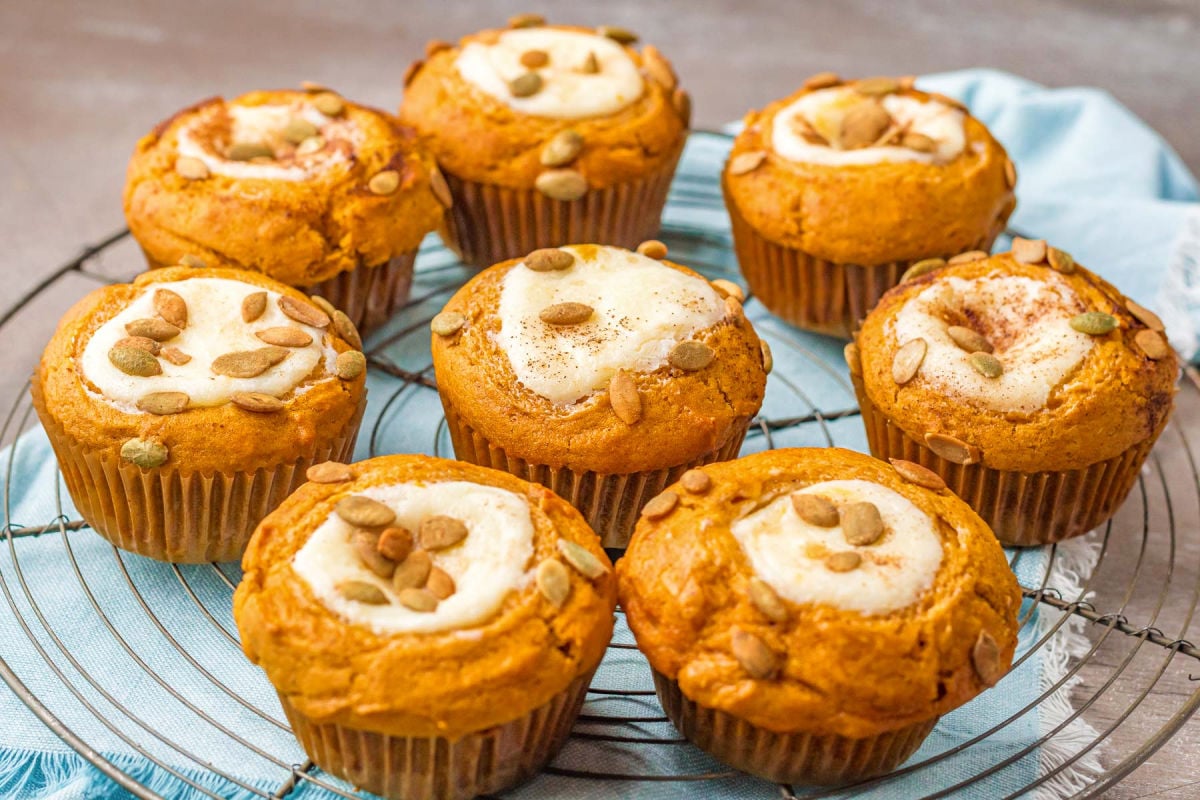 eight pumpkin muffins with cream cheese fillings are sitting on a wire rack on top of a light aqua colored napkin.