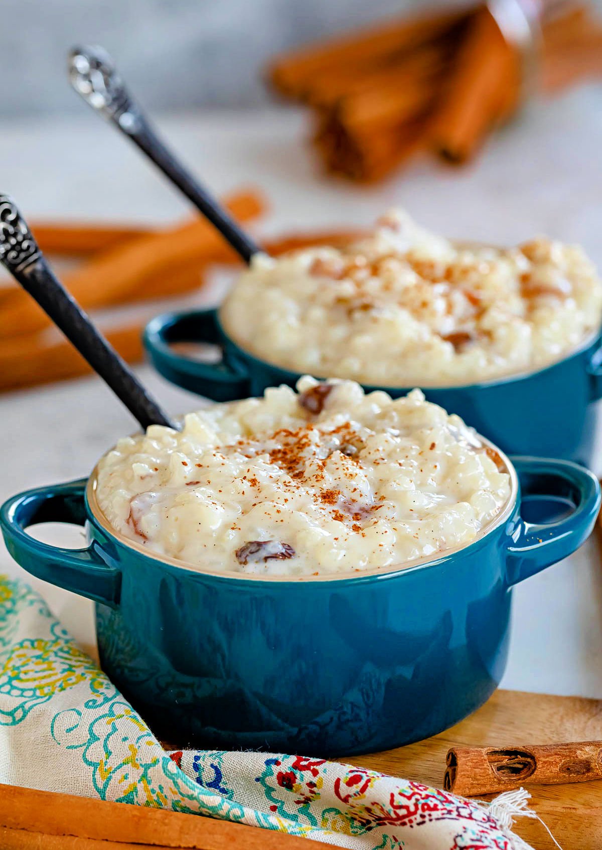 two small bowls filled with rice pudding and both have a cinnamon stick sticking out of the top. garnished with freshly grated cinnamon. each bowl has a black spoon in it,