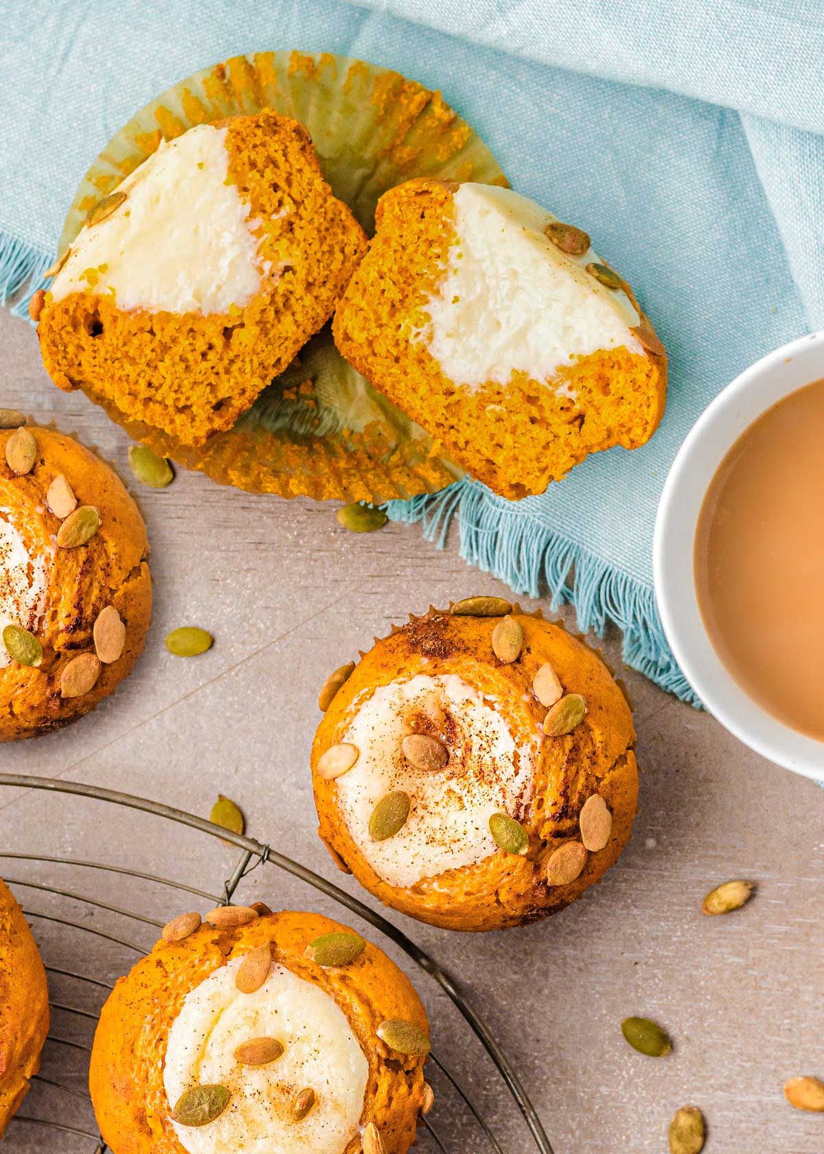 pumpkin cream cheese muffins topped with pumpkin seeds sitting on wood surface with aqua napkin. one muffin has been cut down the middle to reveal the luscious cream cheese fililng.