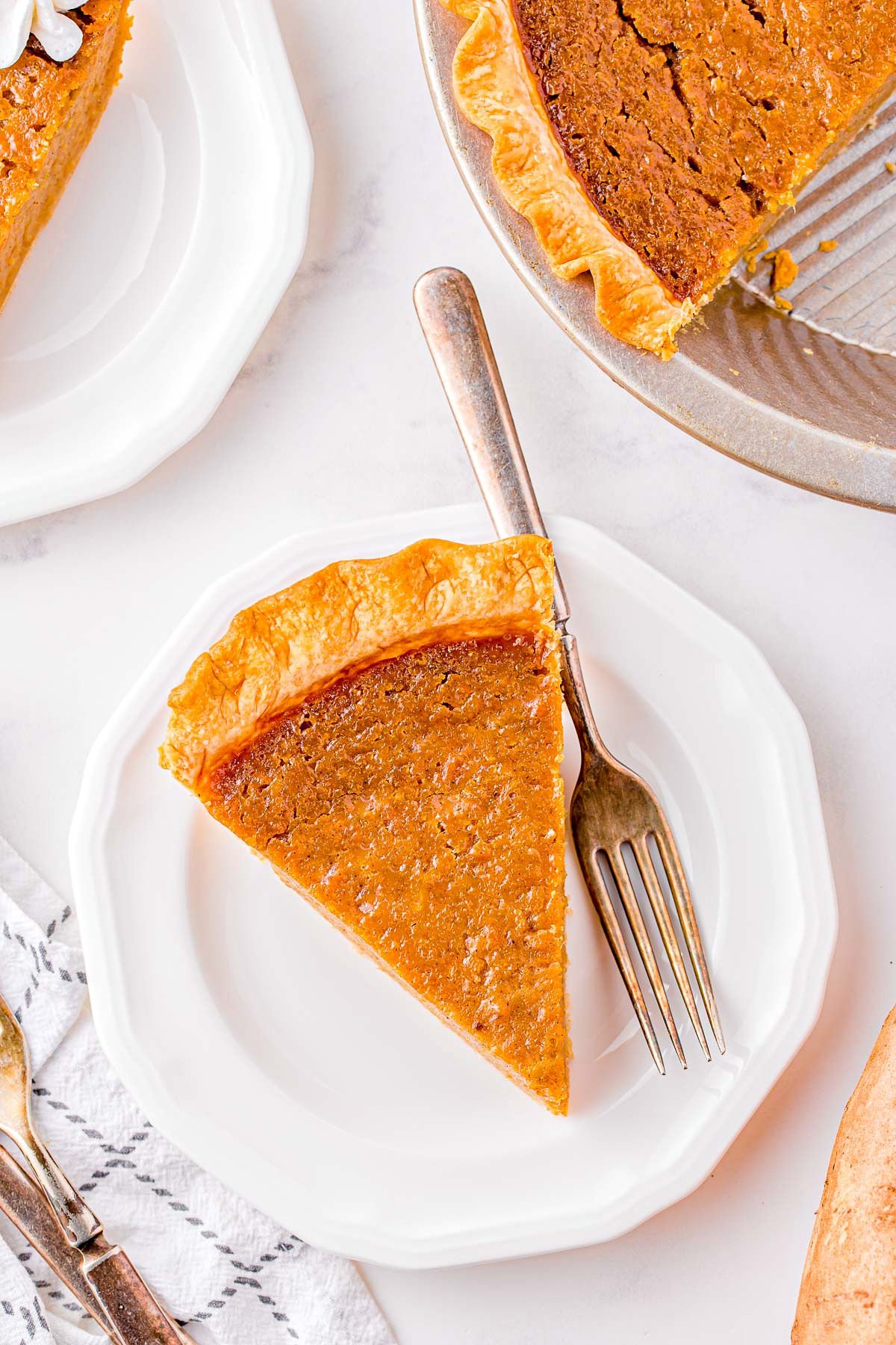 top down look at one piece of sweet potato pie on a white plate with a fork. The rest of the pie and part of another slice are off to the side.