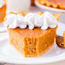 slice of sweet potato pie on white plate topped with whipped cream. the rest of the pie is in the background.