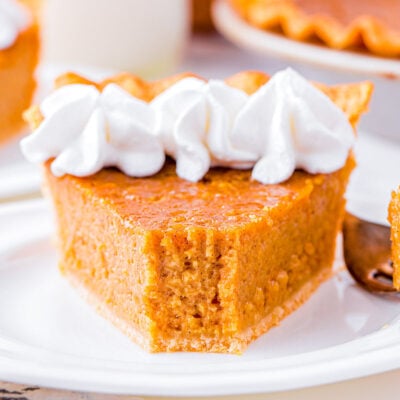 slice of sweet potato pie on white plate topped with whipped cream. the rest of the pie is in the background.