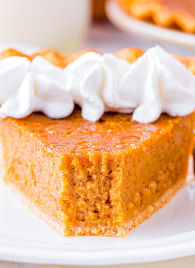 slice of sweet potato pie on white plate topped with whipped cream. the rest of the pie is in the background.