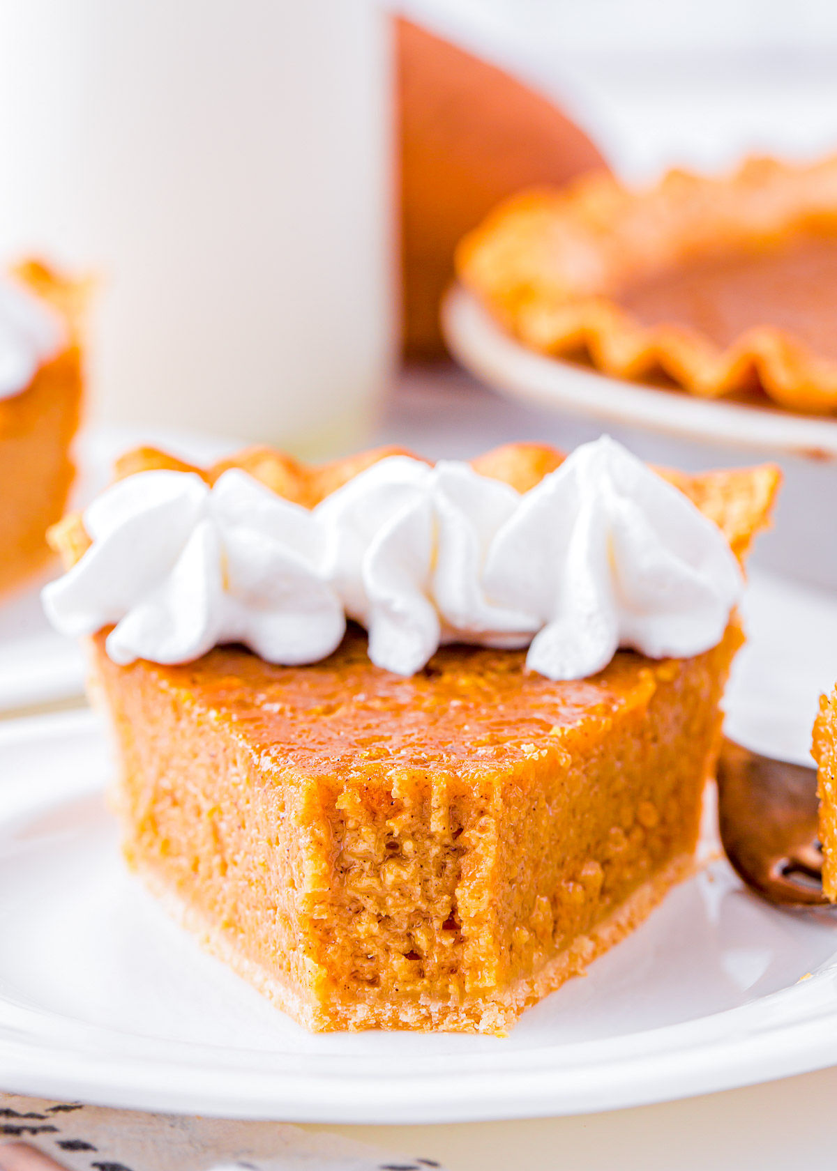 slice of sweet potato pie on white plate topped with whipped cream. the rest of the pie is in the background.