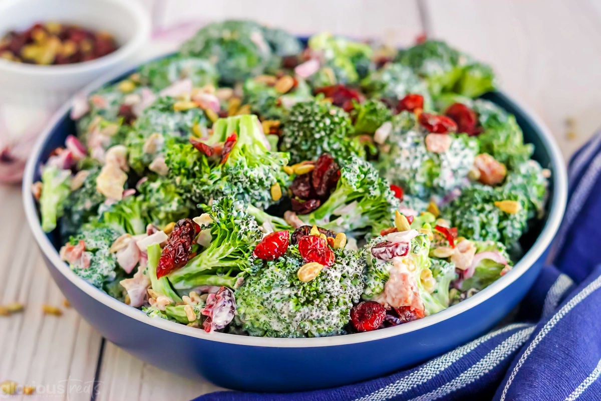 blue bowl with broccoli bacon salad in it ready to be served. blue napkin nestled up against the base of the serving bowl.