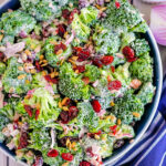 top down look at a broccoli salad made with bacon, cranberries and sunflower seeds in a blue bowl with a blue striped napkin next to it.