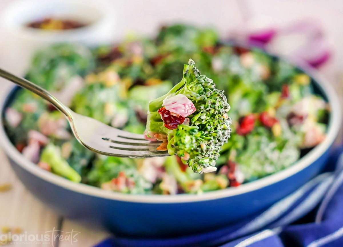 forkful of broccoli salad held in front of a large blue serving bowl with the rest of the broccoli salad recipe.