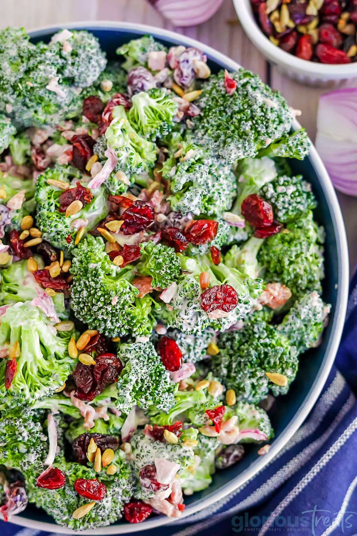 top down look at a broccoli salad made with bacon, cranberries and sunflower seeds in a blue bowl with a blue striped napkin next to it,