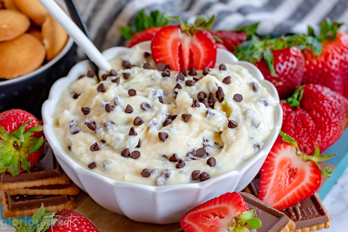 wide shot of cannoli dip in scalloped edge white bowl topped with extra chocolate chips and half a strawberry and spoon stuck in the dip. more strawberries, nilla wafers and cookies are scattered around the bowl as dippers.