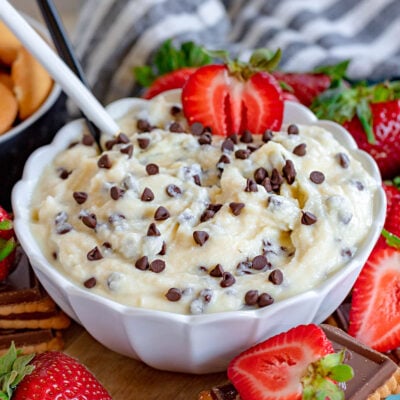 cannoli dip in scalloped edge white bowl topped with extra chocolate chips and half a strawberry and spoon stuck in the dip. more strawberries, nilla wafers and cookies are scattered around the bowl as dippers.