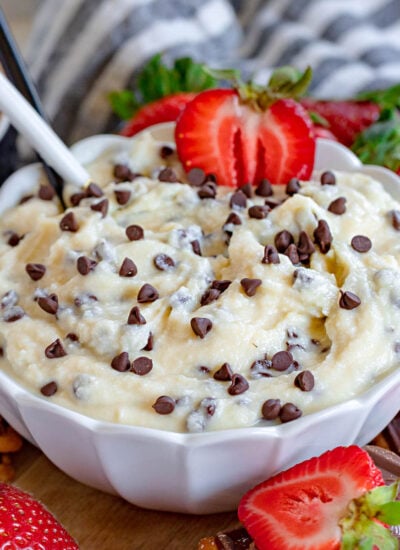 cannoli dip in scalloped edge white bowl topped with extra chocolate chips and half a strawberry and spoon stuck in the dip. more strawberries, nilla wafers and cookies are scattered around the bowl as dippers.