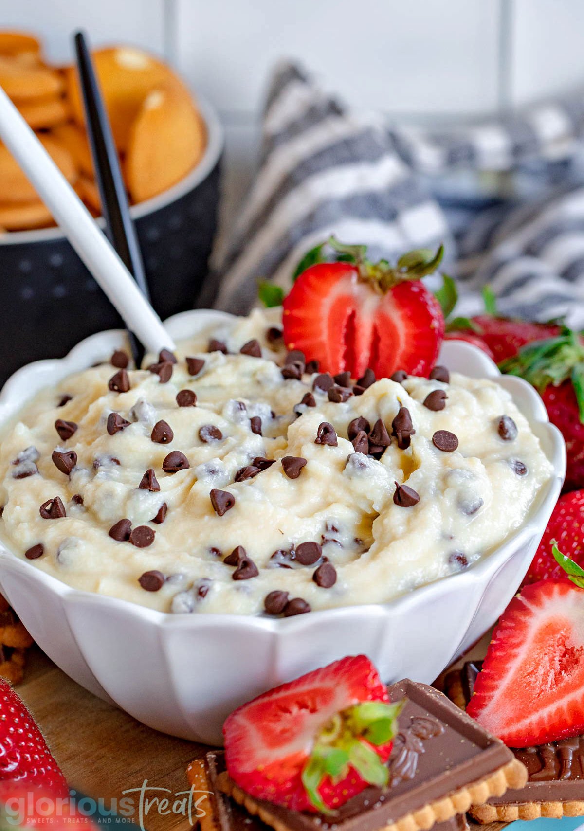 white bowl with scalloped edges filled with cannoli dip and topped with mini chocolate chips. Fresh strawberries, nilla wafers and cookie scattered around the bowl for dipping. white and black spoon inserted into dip.
