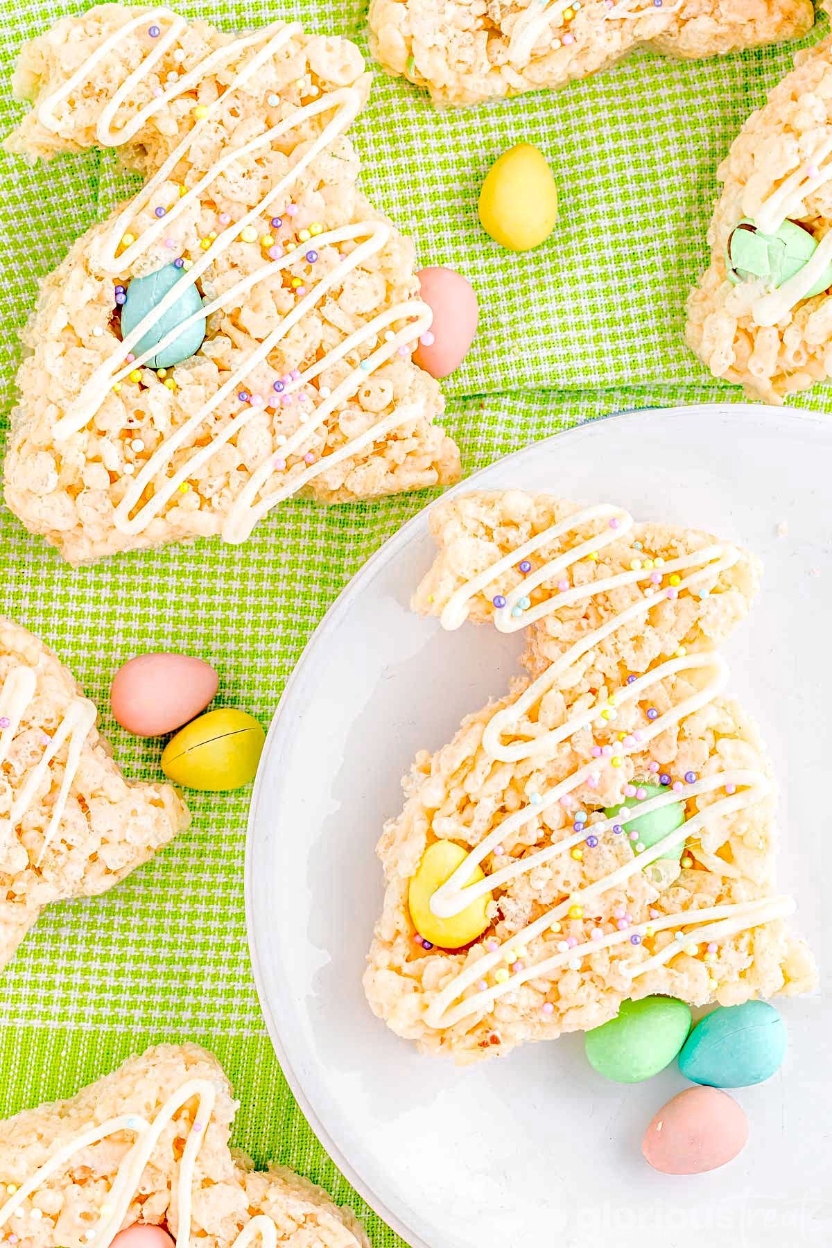 top down look at bunny rice krispie treats topped with a white chocolate drizzle and sprinkles sitting on a green checkered napkin and white plate.