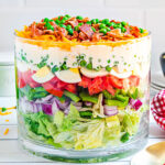 7 layer salad prepared in a clear trifle bowl with straight edges. the salad is topped with more cheese, bacon and green peas. red and white checked napkin to the side.