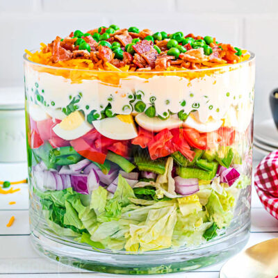 7 layer salad prepared in a clear trifle bowl with straight edges. the salad is topped with more cheese, bacon and green peas. red and white checked napkin to the side.