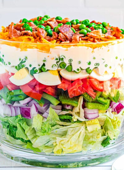 7 layer salad prepared in a clear trifle bowl with straight edges. the salad is topped with more cheese, bacon and green peas. red and white checked napkin to the side.