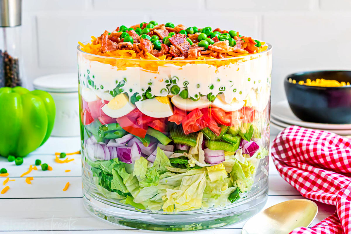 7 layer salad prepared in a clear trifle bowl with straight edges. the salad is topped with more cheese, bacon and green peas. red and white checked napkin to the side.