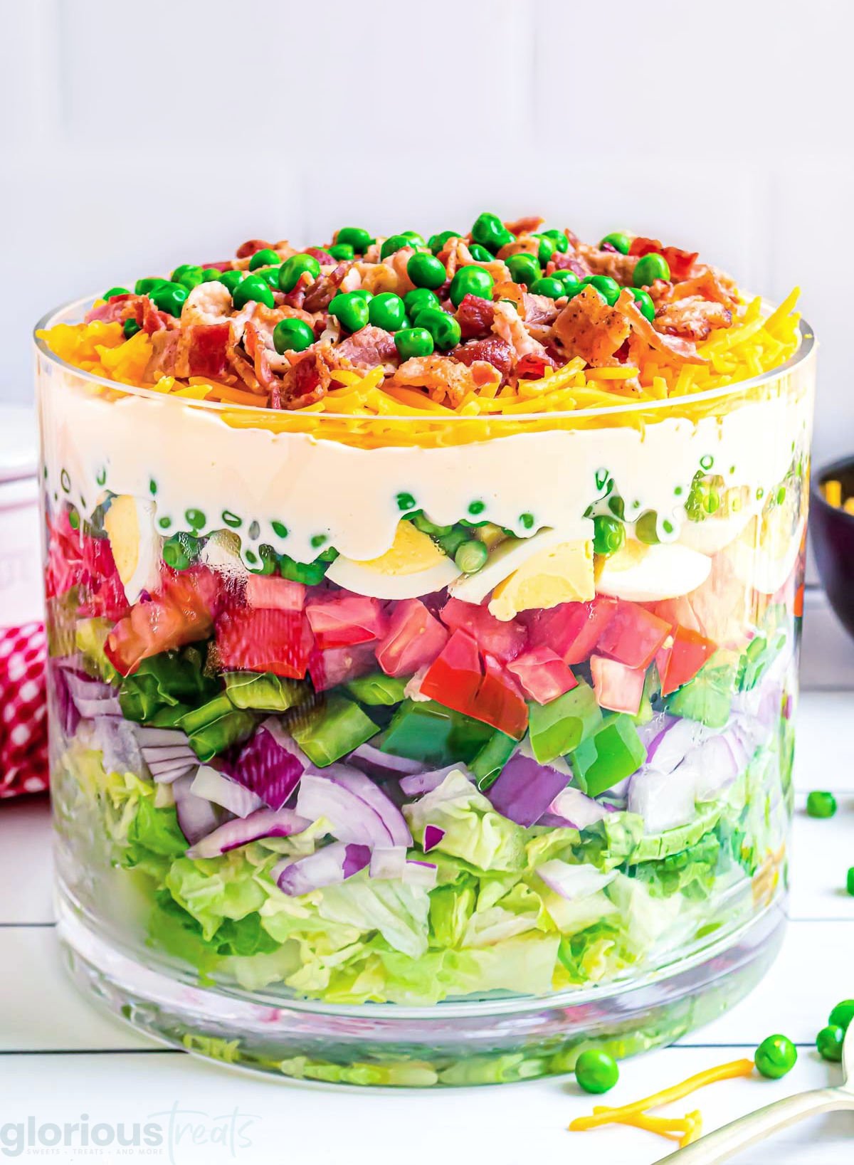seven layer salad prepared in a clear trifle bowl with straight edges. the salad is topped with more cheese, bacon and green peas.