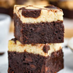 two brownies stacked on top of each other on a small white plate. more brownies can be seen in the background.