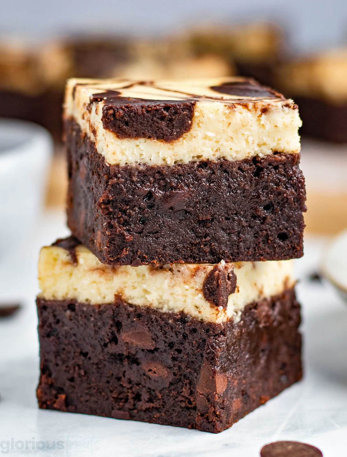 two brownies stacked on top of each other on a small white plate. more brownies can be seen in the background.