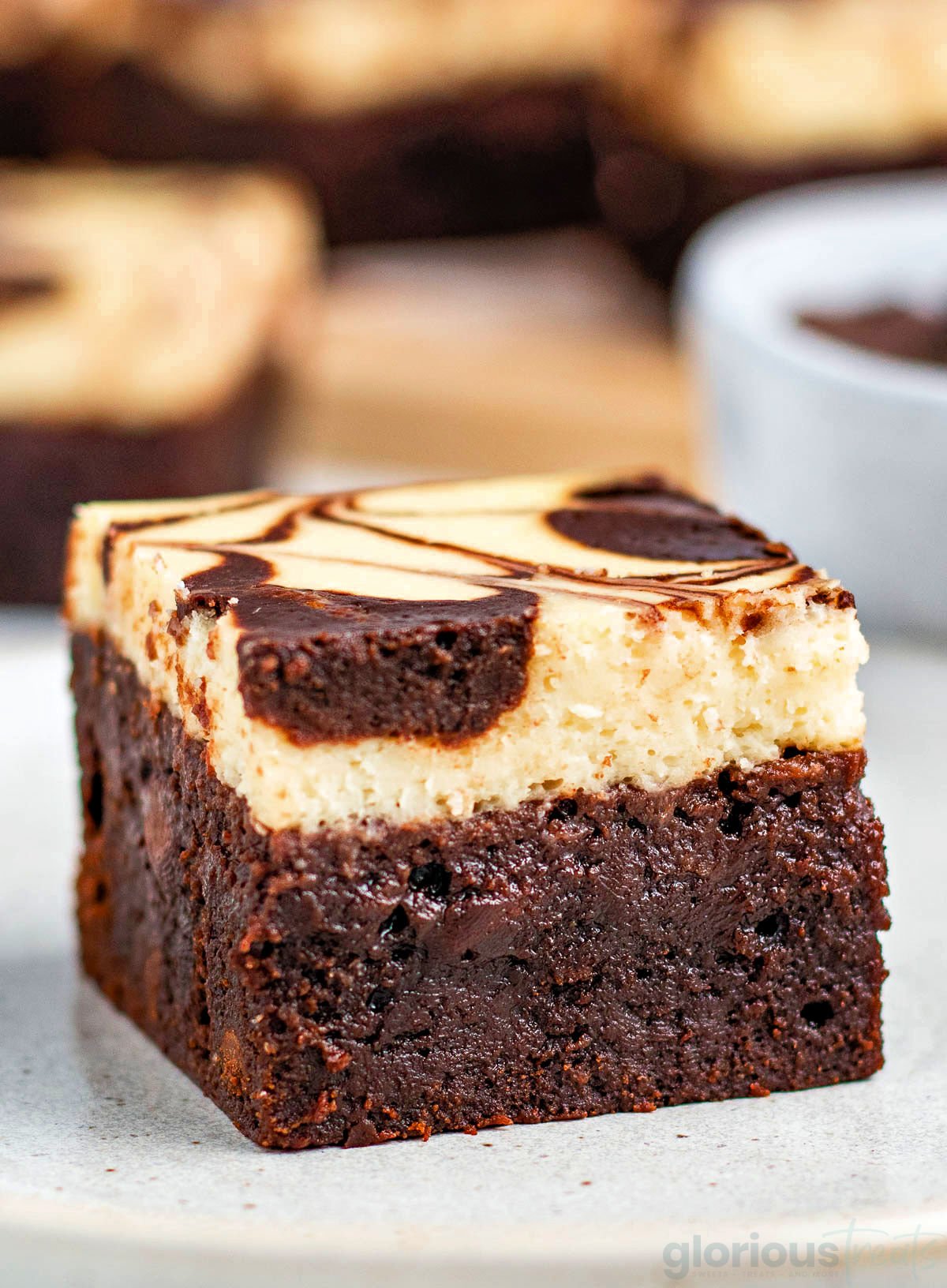 a single cheesecake brownie sitting on a small white plate with more brownies in the background.