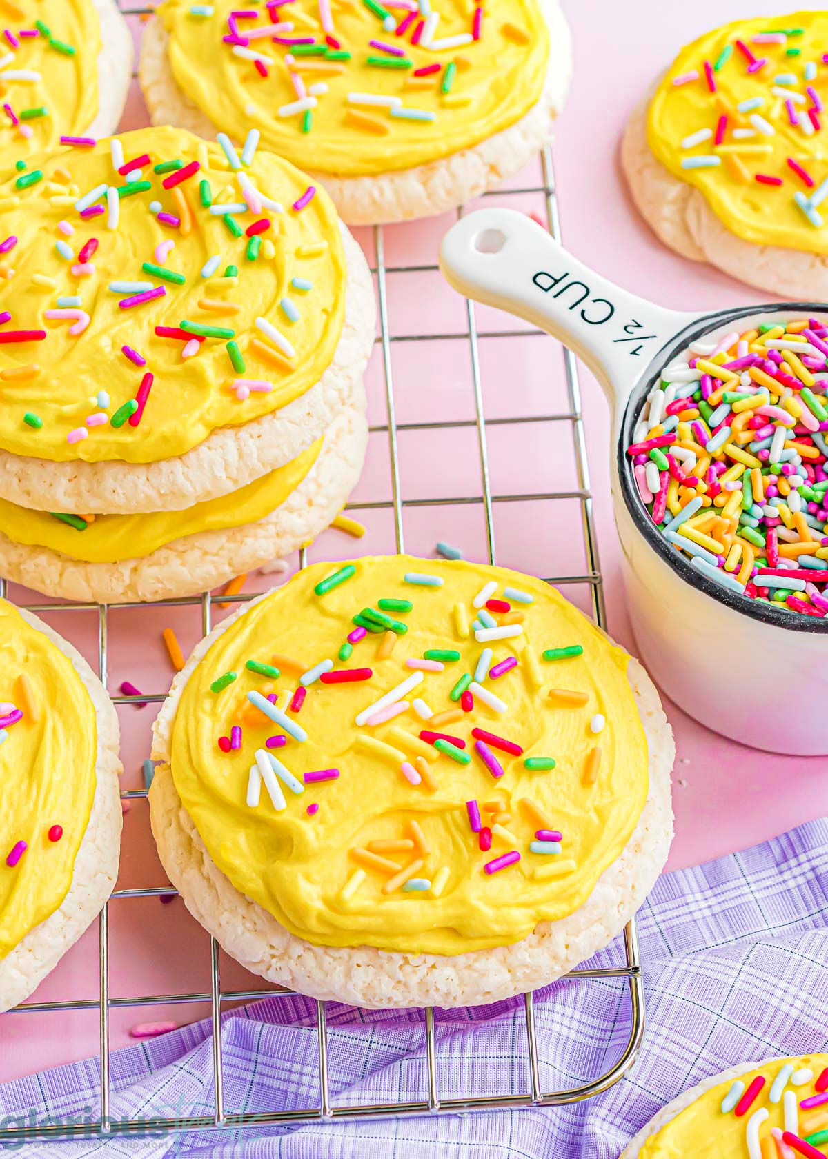 lots of lofthouse sugar cookies on a cooling rack that have been frosting with yellow frosting. Rainbow sprinkles are scattered about and top each cookie. 