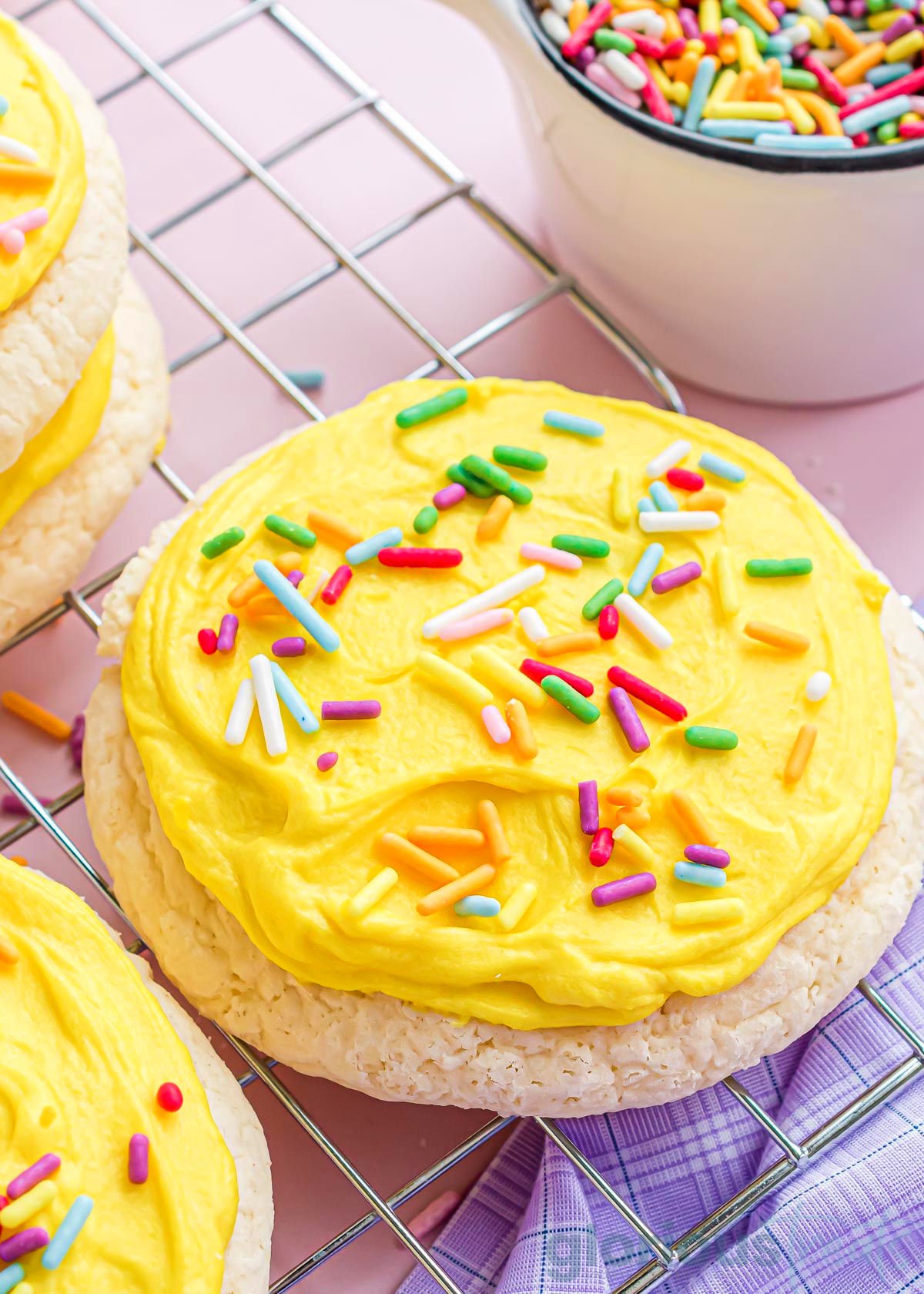 a cooling rack sitting on a pink surface with a lavender kitchen towel topped with lots of lofthouse cookies. the cookies have yellow frosting and rainbow jimmies.