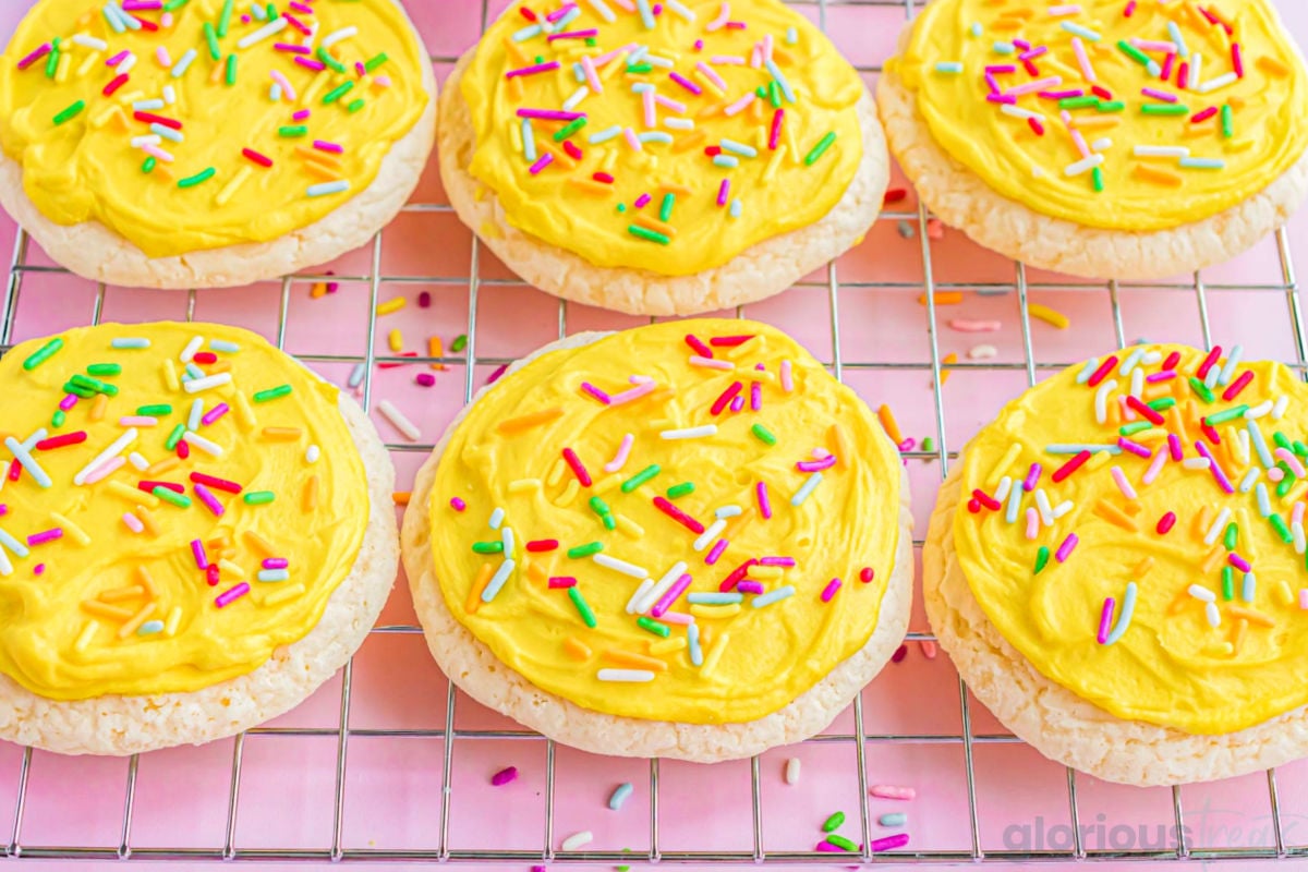 six lofthouse cookies sitting on a cooling rack sitting on a pink surface. cookies are frosted with yellow frosting and rainbow sprinkles.