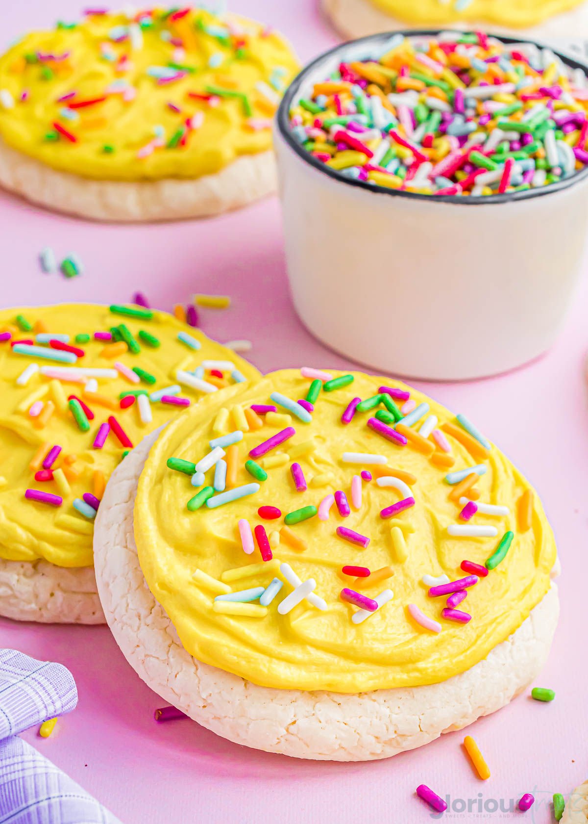 lofthouse cookies topped with bright yellow frosting and colorful jimmies. a small measuring cup filled with more sprinkles in the background.