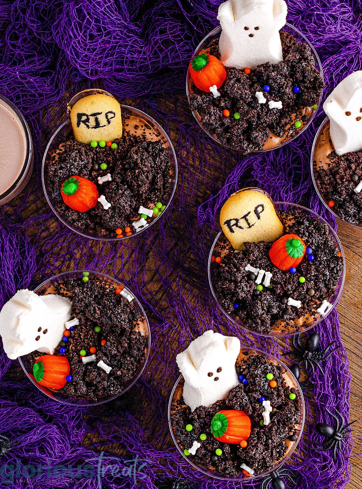 top down look at halloween graveyard dirt cups decorated with halloween candy and make with oreos and chocolate pudding. the cups are sitting on a bright purple net.