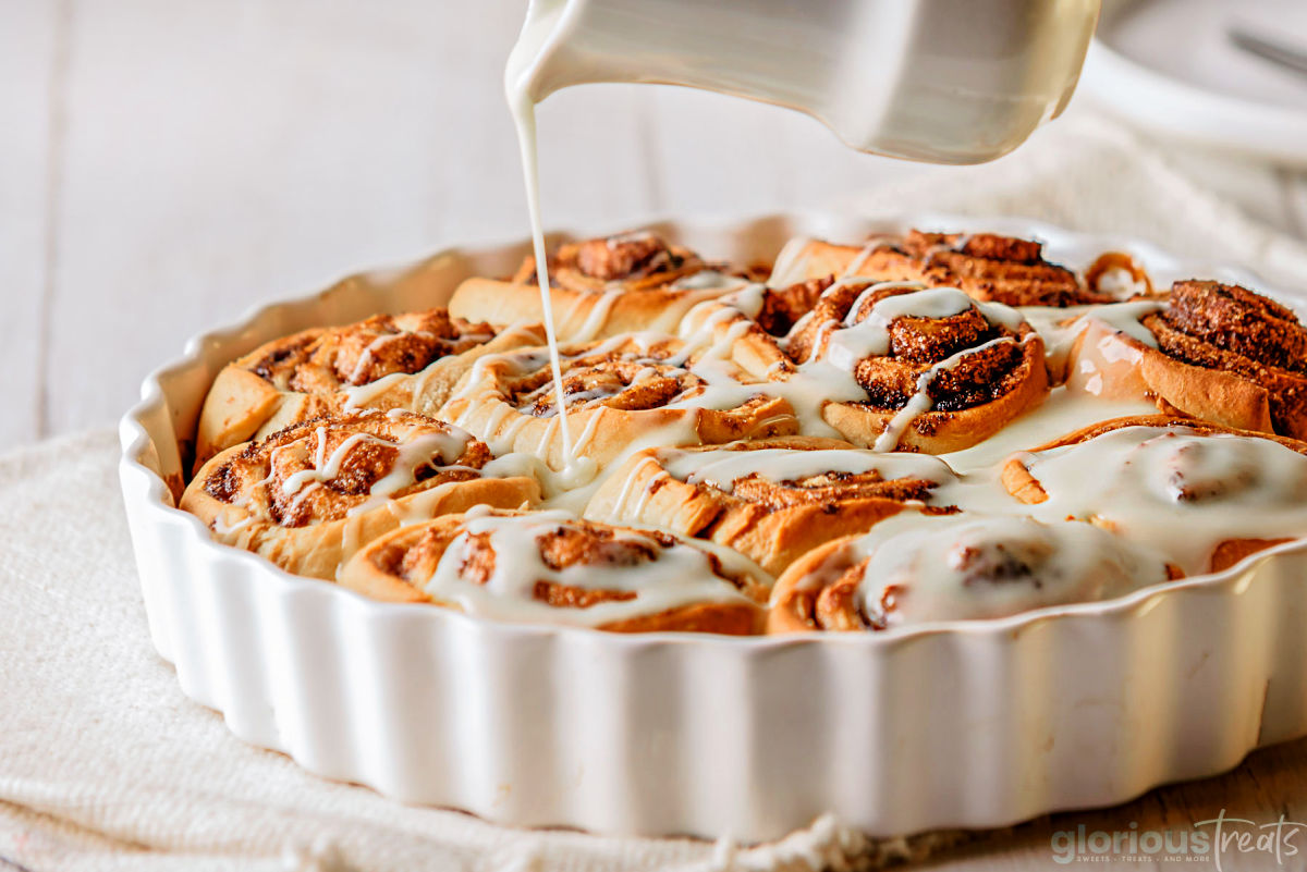 cinnamon roll icing being drizzle over fresh baked cinnamon rolls in a white baking dish.
