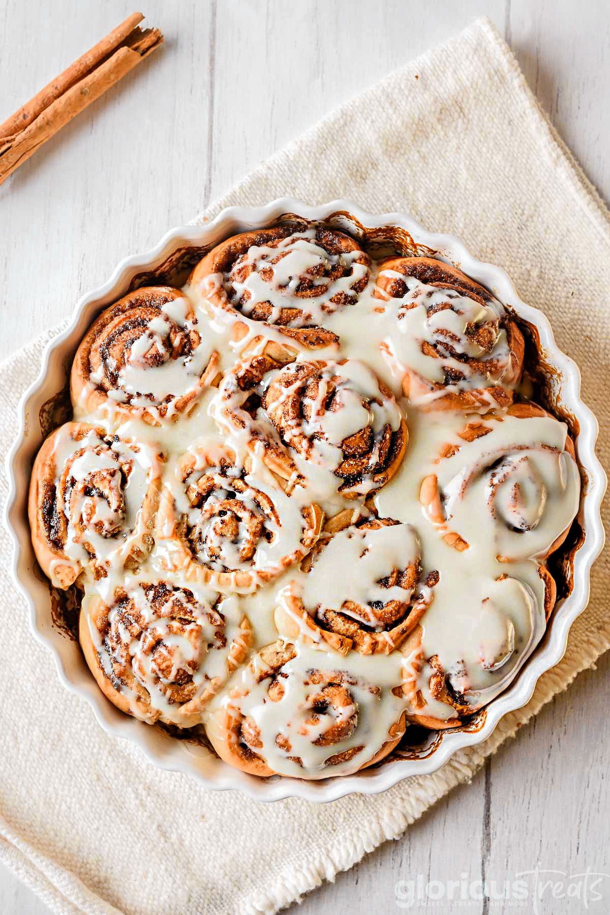 top down look at homemade cinnamon rolls topped with icing in a round white scalloped casserole dish with a beige napkin beneath.