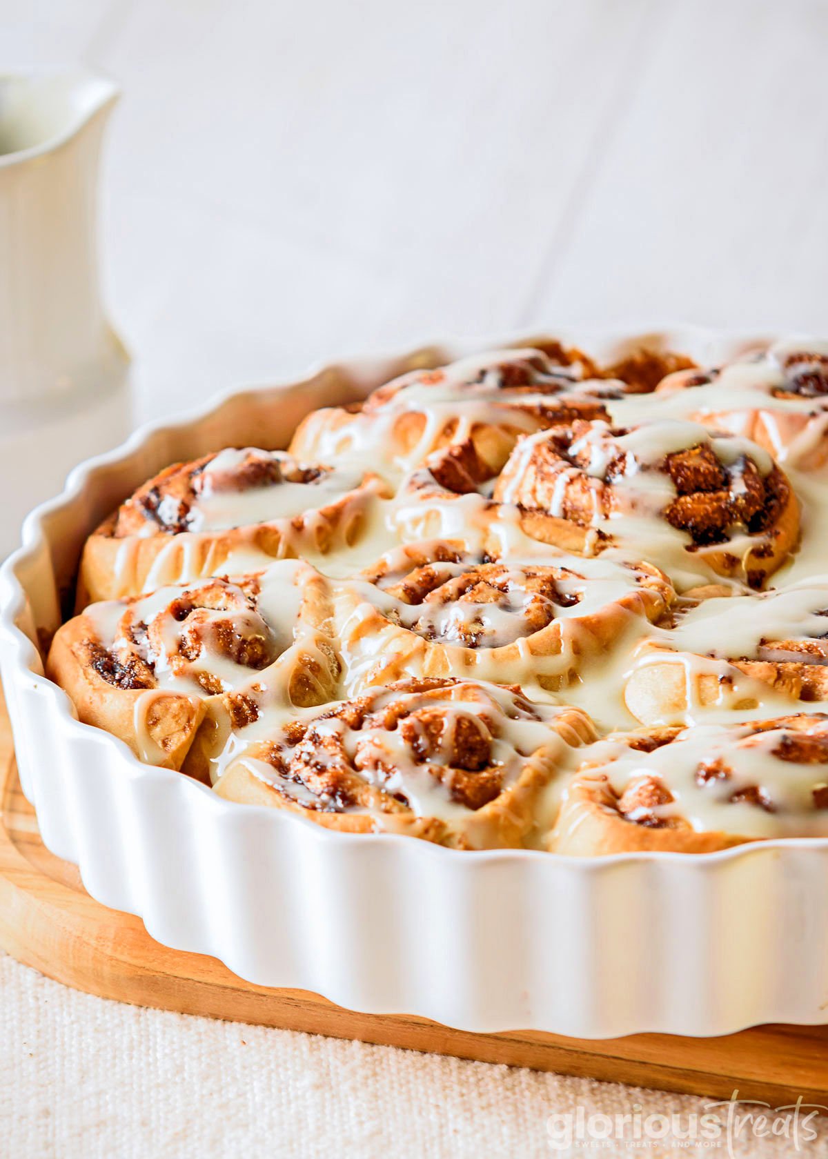 cinnamon rolls recipe topped with cream cheese cinnamon roll icing in a white round baking dish. baking dish is sitting on wood board.