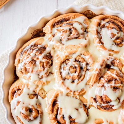 top down look at homemade cinnamon rolls topped with icing in a round white scalloped casserole dish with a beige napkin beneath.