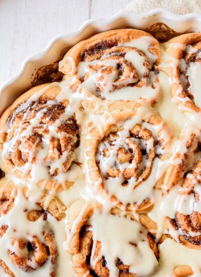 top down look at homemade cinnamon rolls topped with icing in a round white scalloped casserole dish with a beige napkin beneath.