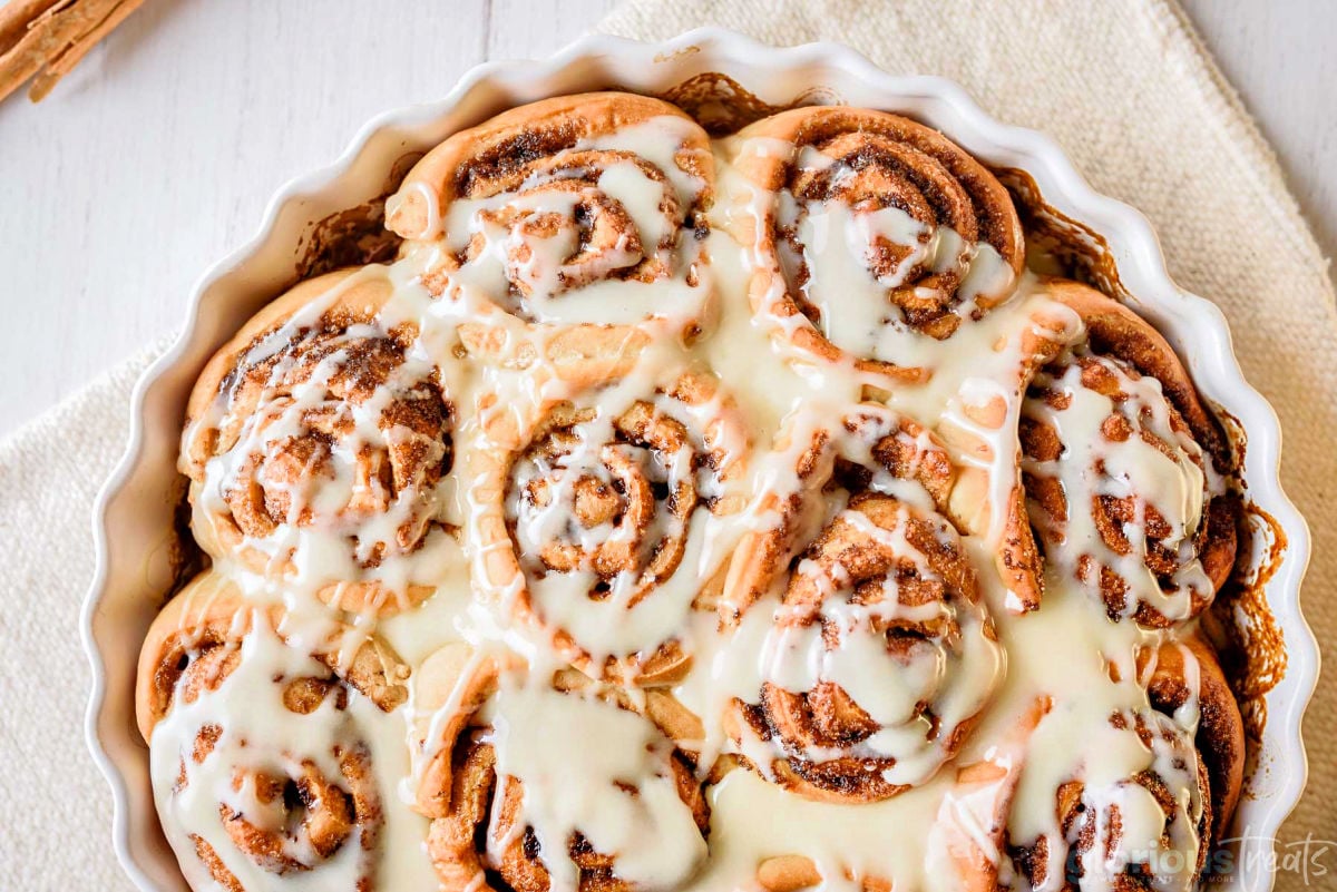 top down look at homemade cinnamon rolls topped with icing in a round white scalloped casserole dish with a beige napkin beneath.