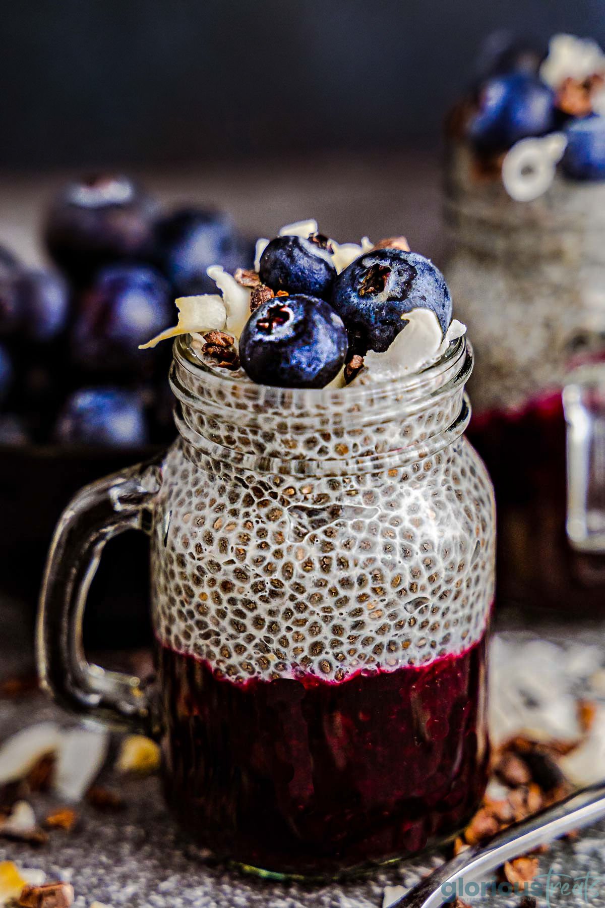 chia seed pudding recipe in a mason jar glass with half a lemon sitting next to it. the pudding is topped with fresh blueberries and coconut flakes and the pudding is layered with blueberry compote.
