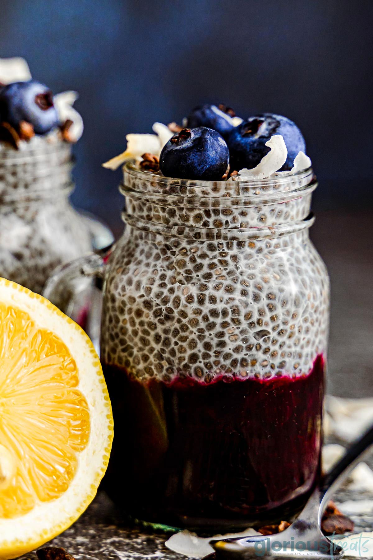 chia pudding in a mason jar glass with half a lemon sitting next to it. the pudding is topped with fresh blueberries and coconut flakes and the pudding is layered with blueberry compote.