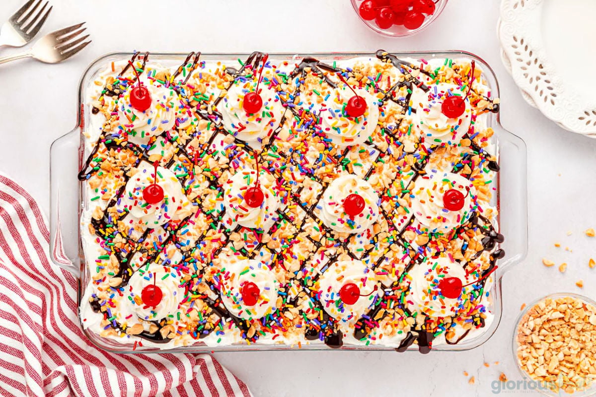 top down look at assembled no bake banana split dessert in a glass baking dish topped with sprinkles and maraschino cherries.
