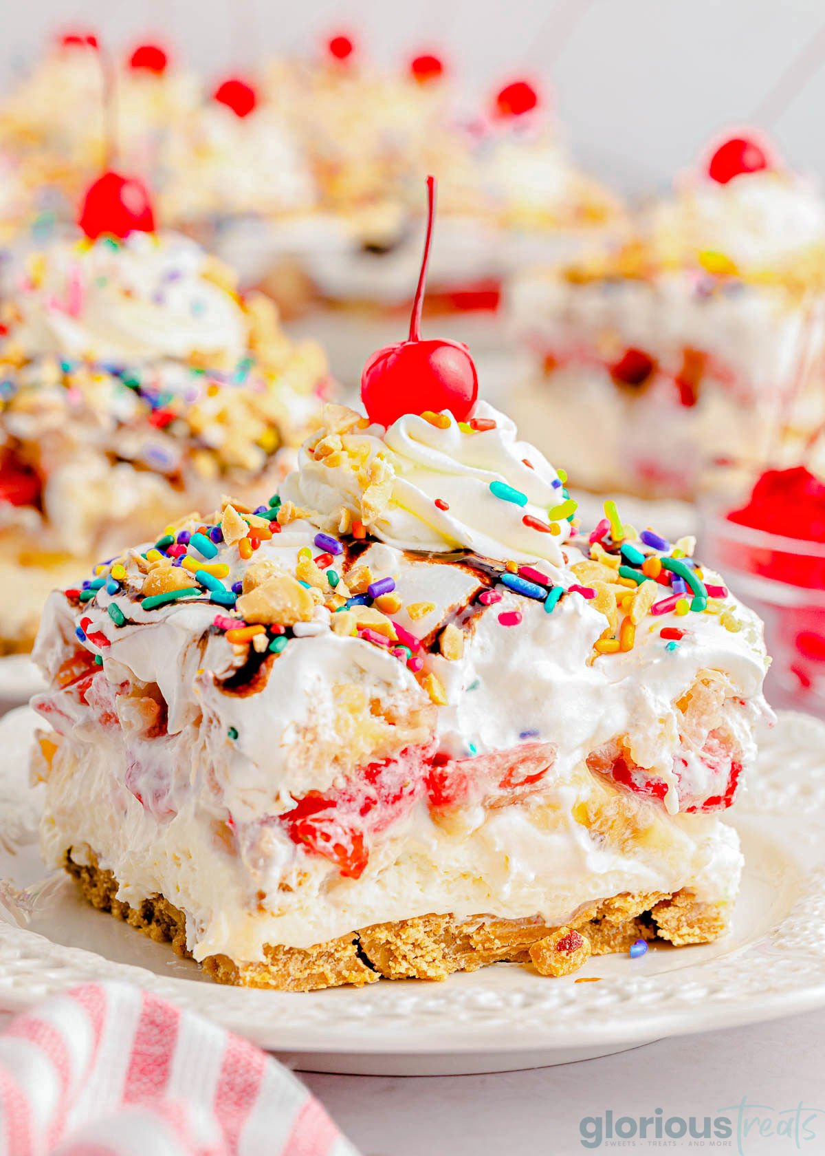 serving of no bake banana split dessert on white plate topped with whipped cream and a maraschino cherry.