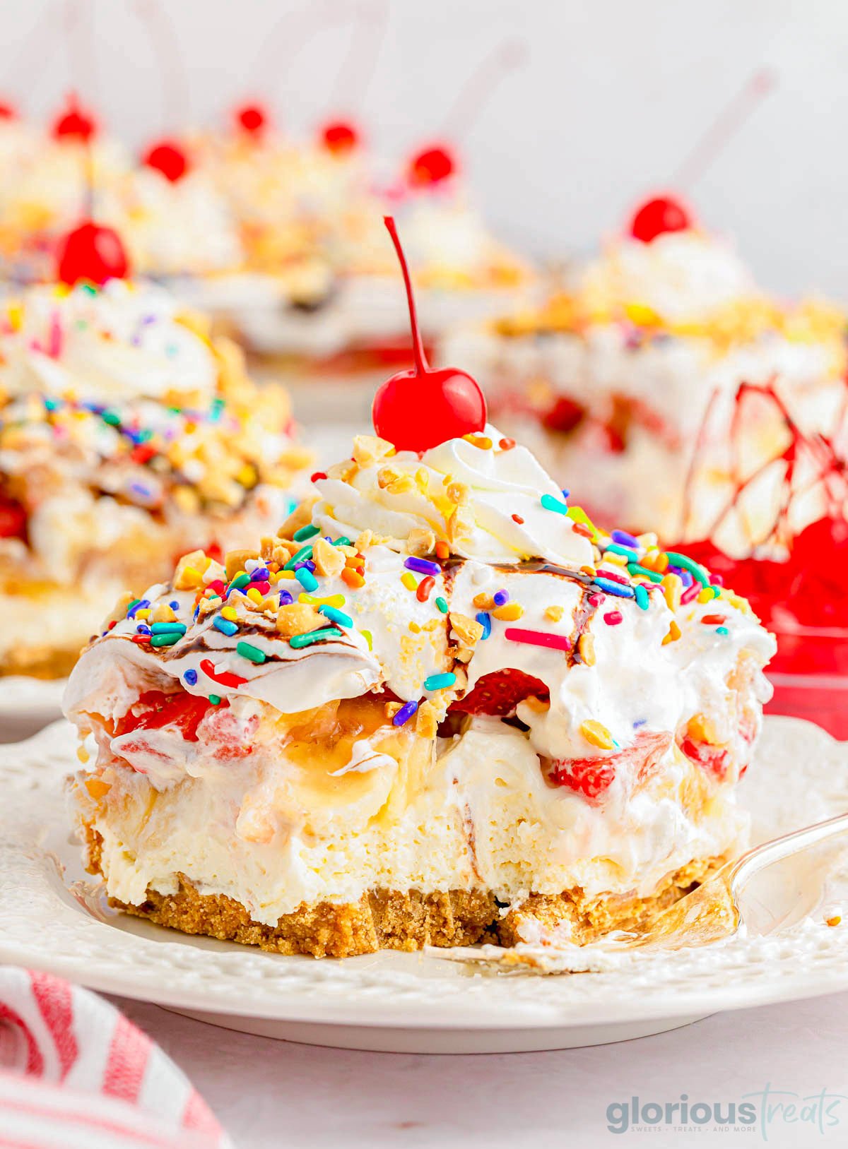 serving of no bake banana split dessert on white plate topped with whipped cream and a maraschino cherry. A bite has been taken out of the dessert.