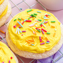 a cooling rack sitting on a pink surface with a lavender kitchen towel topped with lots of lofthouse cookies. the cookies have yellow frosting and rainbow jimmies.