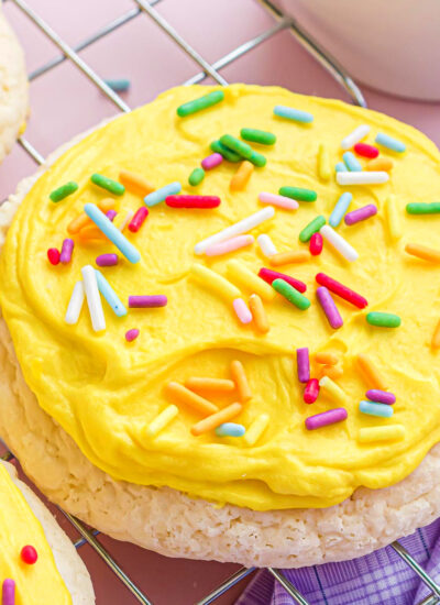 a cooling rack sitting on a pink surface with a lavender kitchen towel topped with lots of lofthouse cookies. the cookies have yellow frosting and rainbow jimmies.